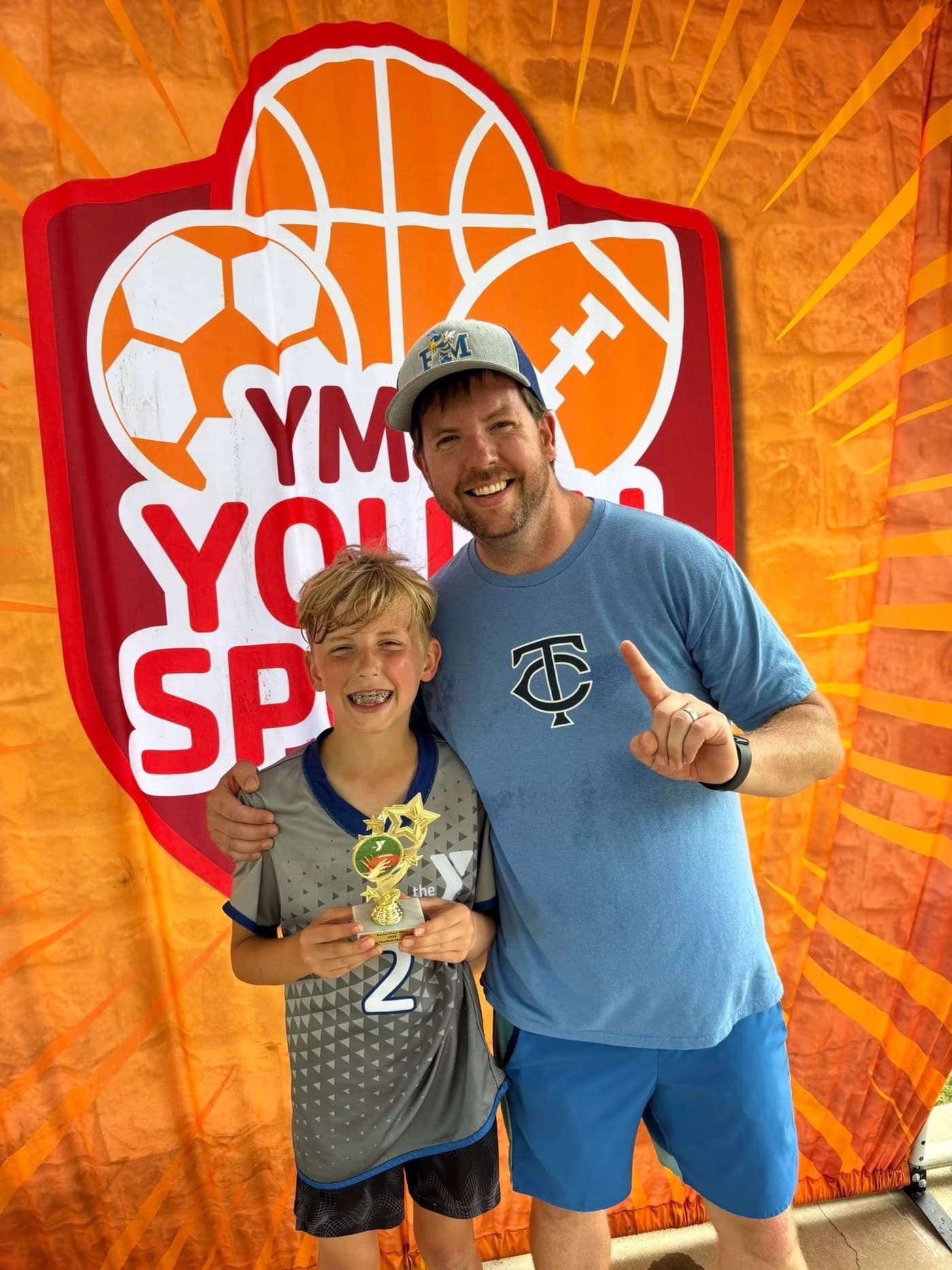 Joe Helms in a Fort Mill cap with his son, who is holding a YMCA trophy, in front of a YMCA Youth Sports banner.