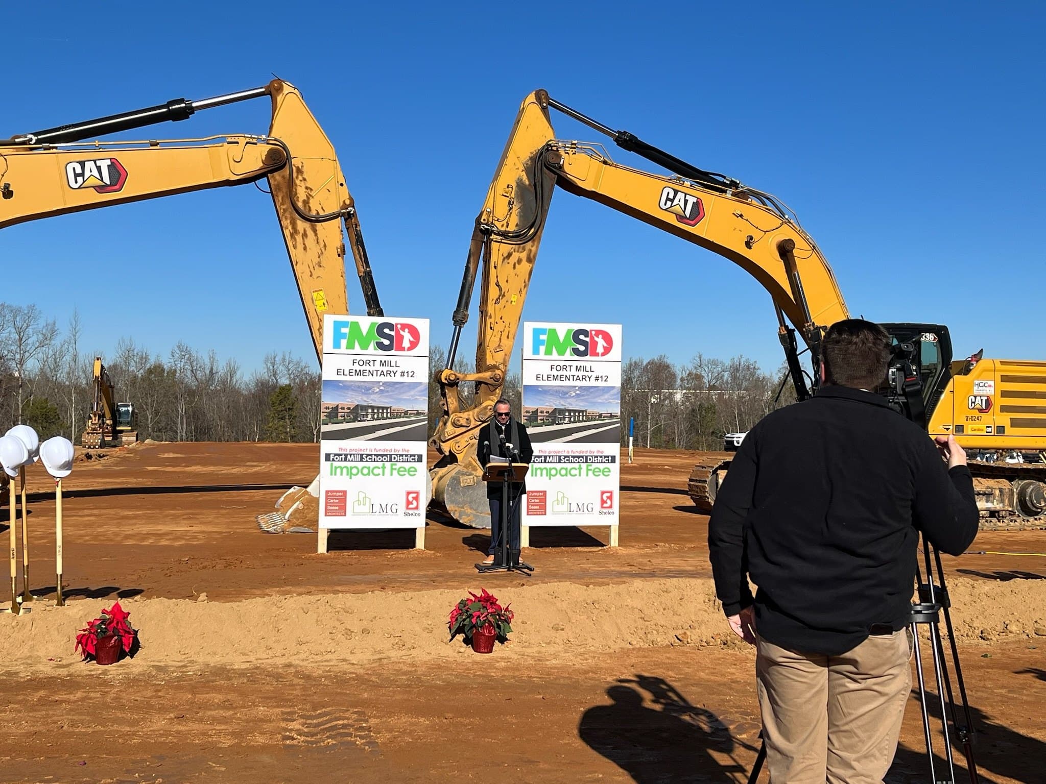 The groundbreaking ceremony for Flint Hill Elementary. Two excavators flank a pair of Fort Mill School District signs reading "Fort Mill Elementary #12", with ceremonial shovels and hard hats in the foreground and a speaker at a podium.