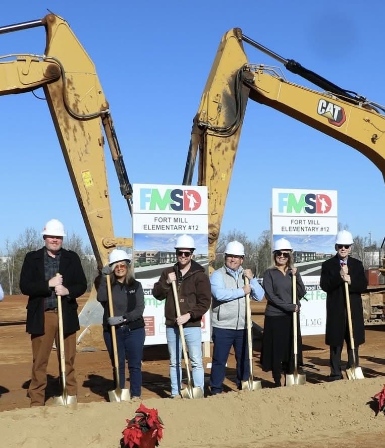 Six people in hard hats holding ceremonial shovels at the Flint Hill groundbreaking, between two excavators and the Fort Mill School District signs.