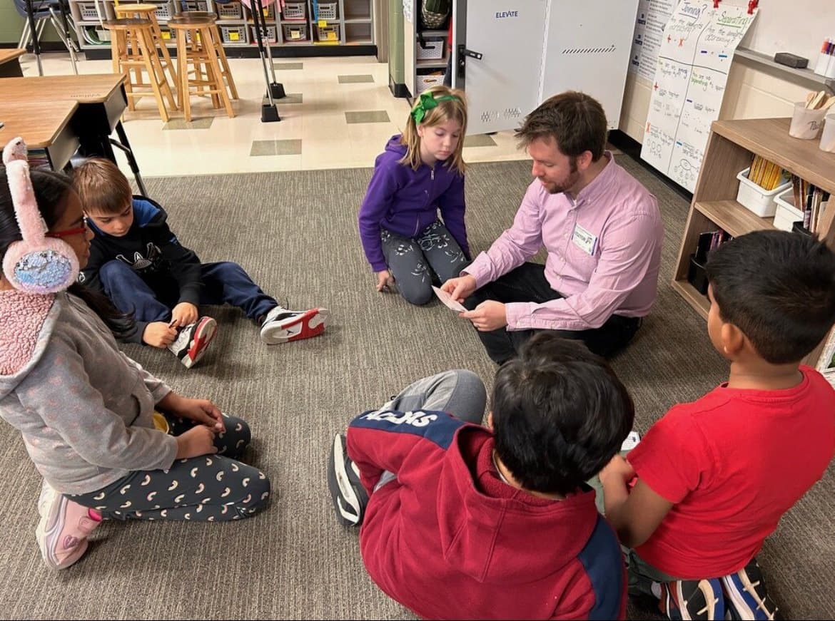 Joe Helms sitting on the floor in a circle with a group of young students in a Fort Mill classroom, holding a set of cards.