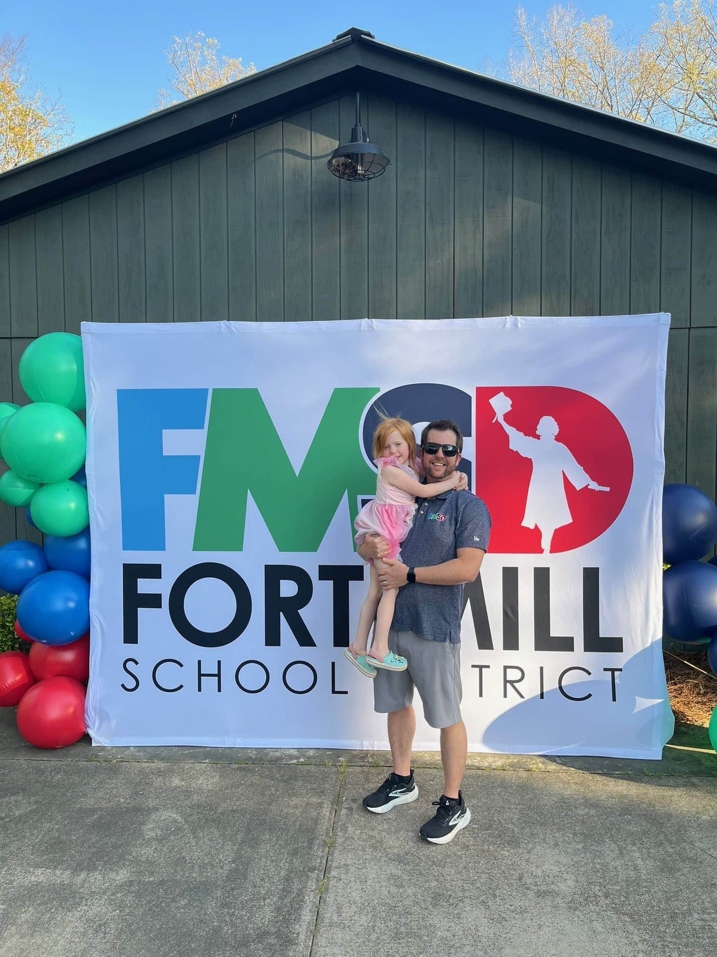 Joe Helms holding his youngest daughter in front of a large Fort Mill School District banner at an outdoor event.