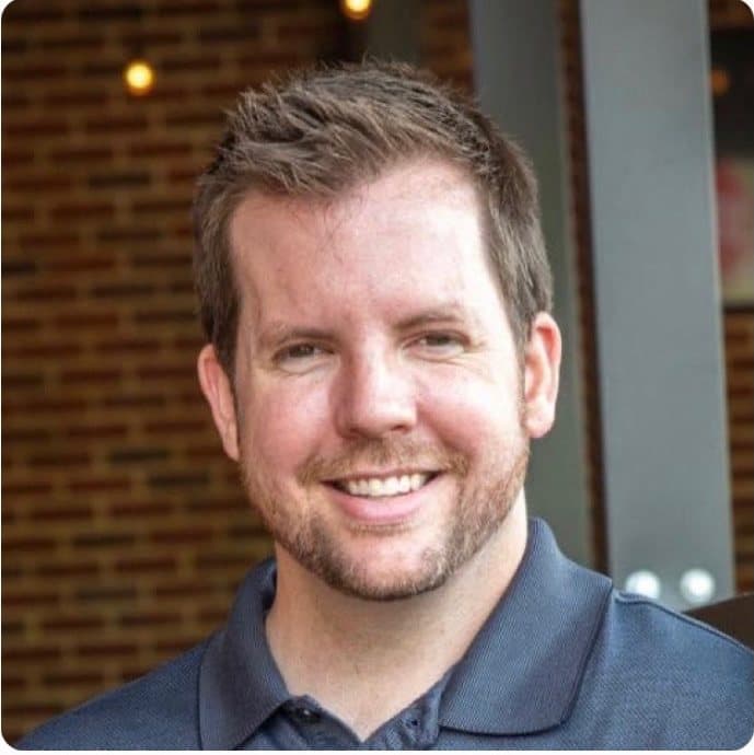 Joe Helms smiling in a navy polo shirt outside a brick building.
