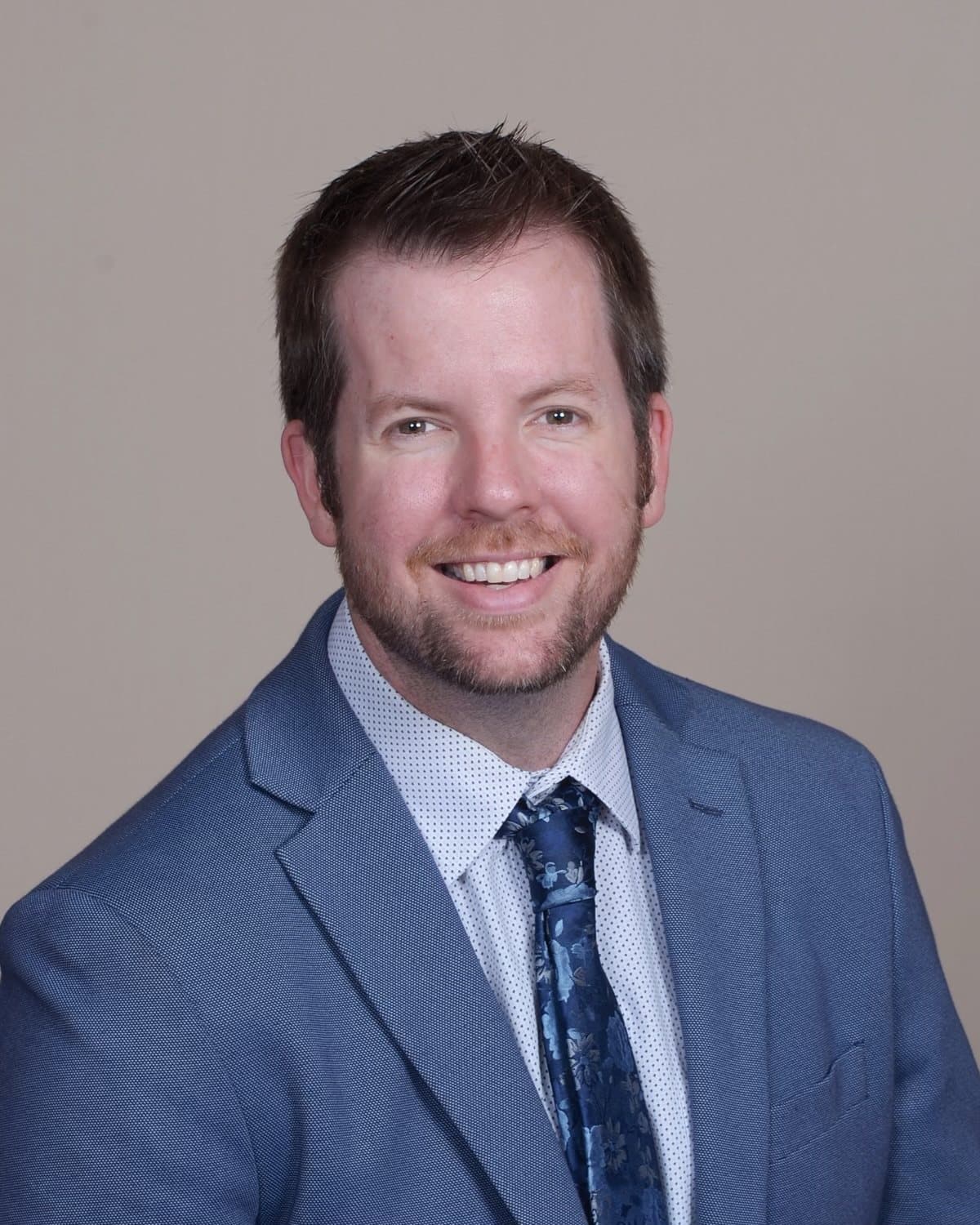 Joe Helms in a blue suit and patterned tie, photographed against a warm neutral backdrop.