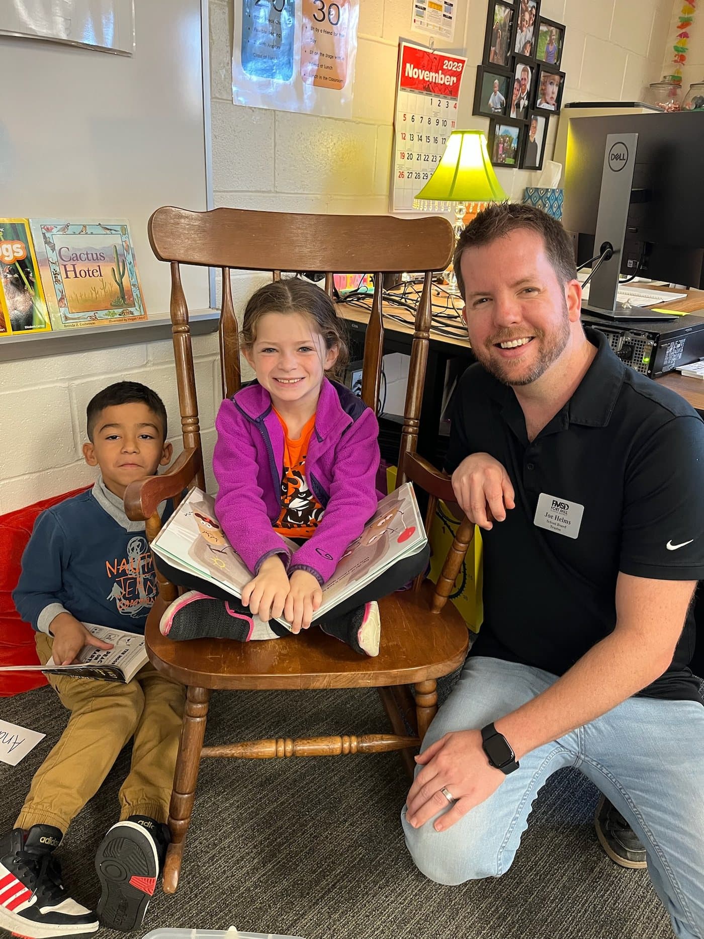 Joe Helms kneeling beside two young students reading a picture book in a Fort Mill classroom, wearing his school board trustee badge.