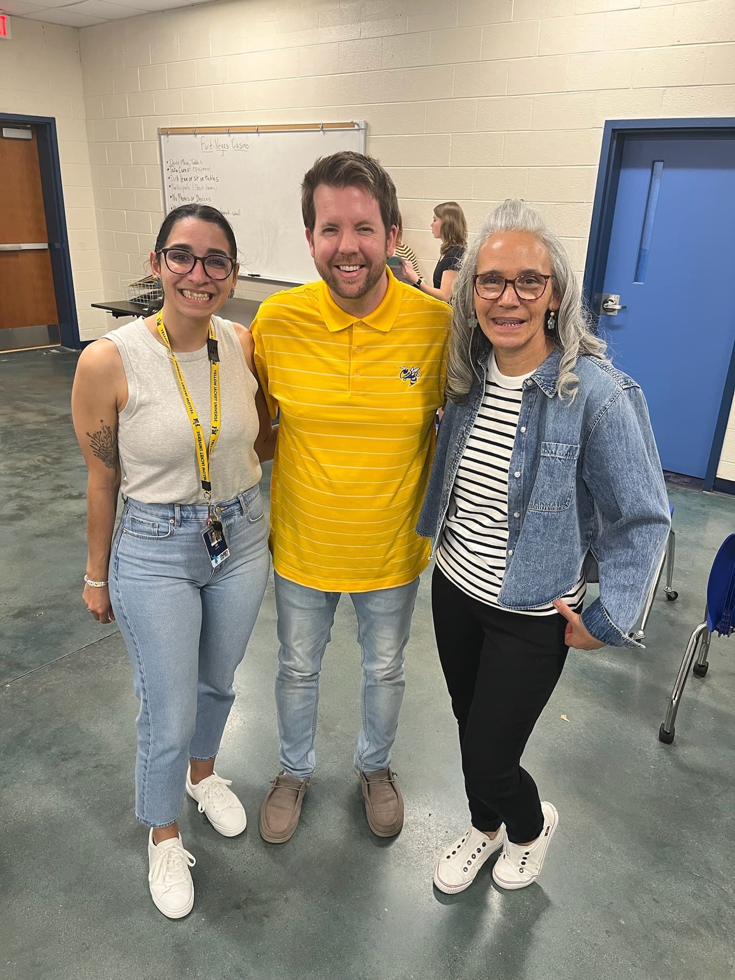 Joe Helms in a yellow shirt standing between two Fort Mill teachers in a school classroom.