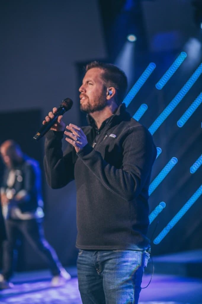 Joe Helms singing into a handheld microphone on a stage lit in blue, leading worship at his church, with a guitarist behind him.